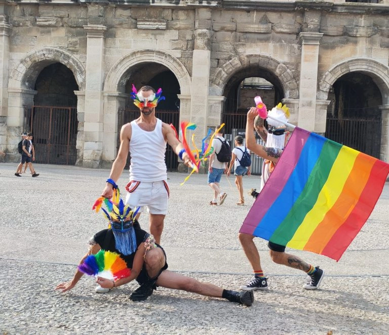 Personnes en costume avec drapeaux arc-en-ciel devant arènes.
